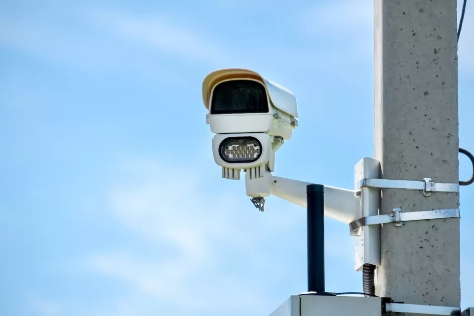 Close-up of white traffic enforcement camera mounted on concrete pole against blue sky.
